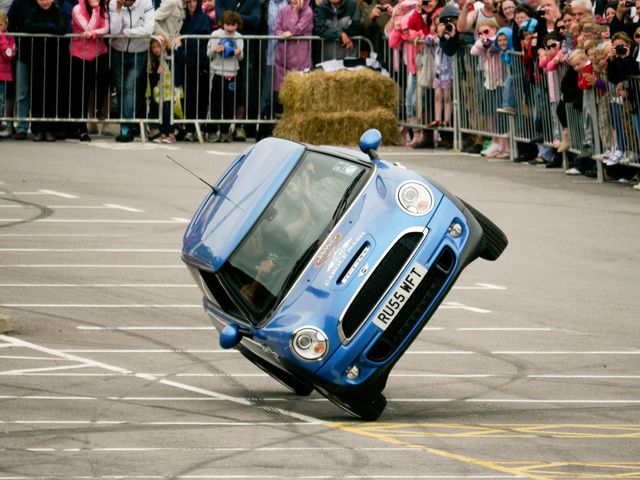 Una Mini Cooper blu in equilibrio su due ruote durante uno spettacolo di stunt driving